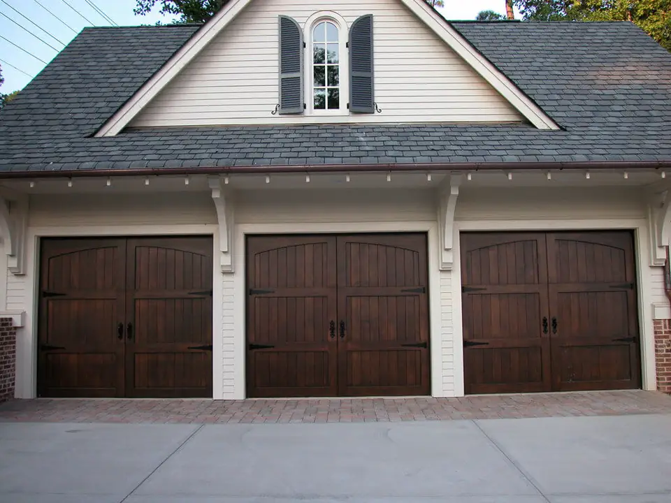 Three dark-stained wood carriage garage doors with arched top panels, black strap hinges and ring handles, on a cream clapboard garage with a dormer.