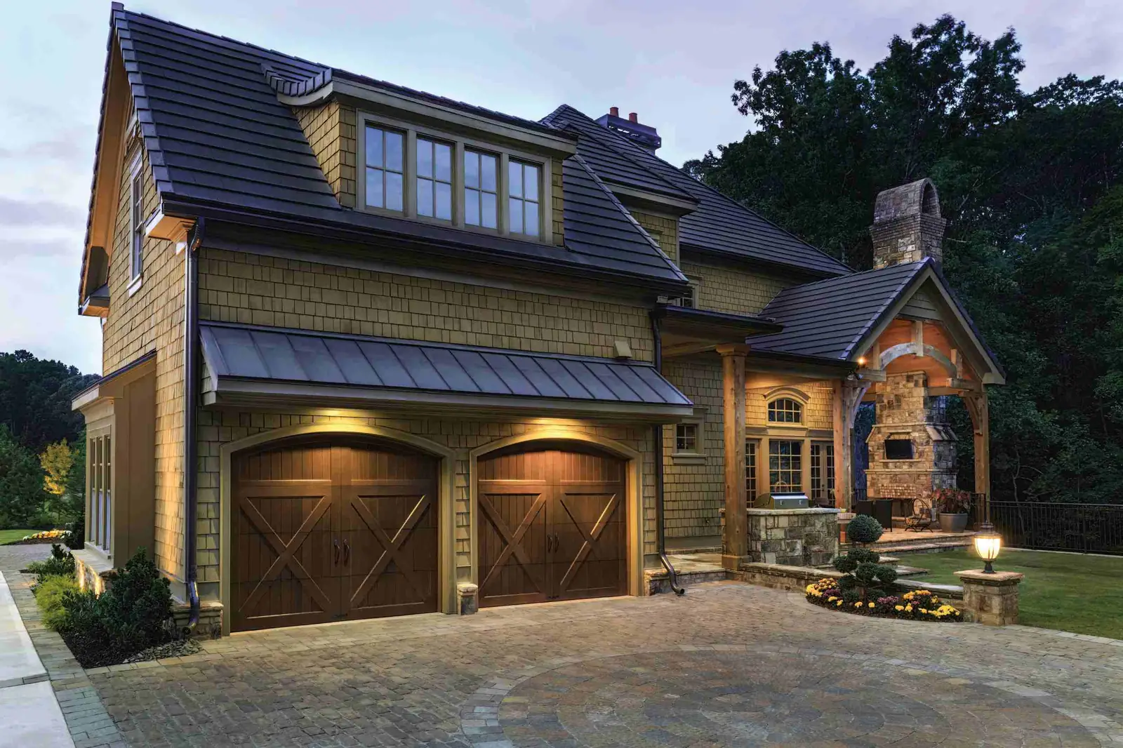 Two arched-top brown carriage garage doors with X-brace panels and no windows, under a metal awning on a shingle-sided house at dusk.