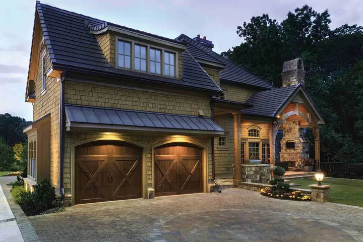 Two arched-top brown carriage garage doors with X-brace panels and no windows, under a metal awning on a shingle-sided house at dusk.
