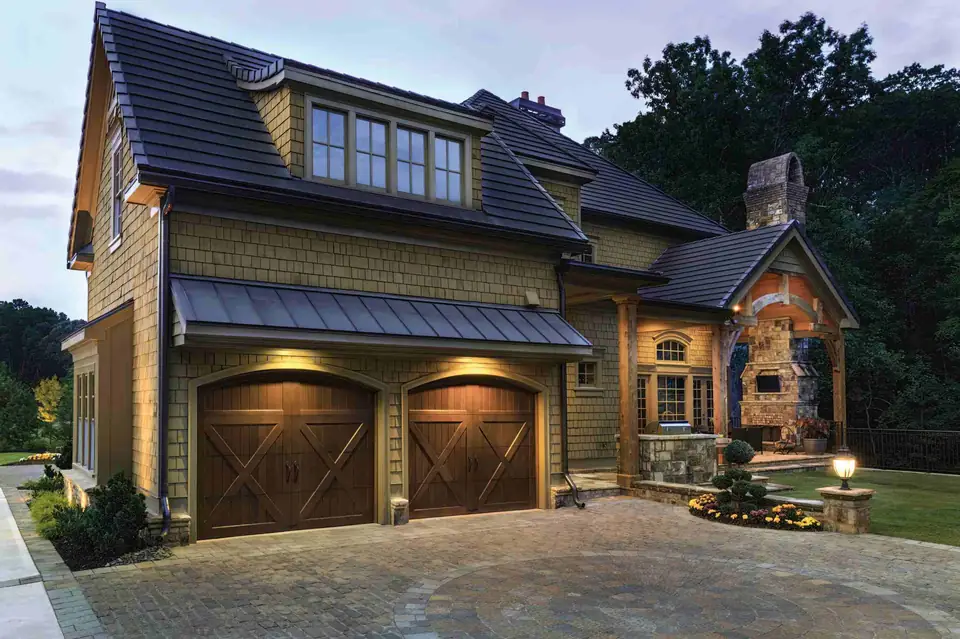 Two arched-top brown carriage garage doors with X-brace panels and no windows, under a metal awning on a shingle-sided house at dusk.