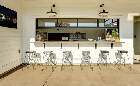 Black-framed aluminum and glass overhead door raised open over an outdoor bar counter, with six wire stools and white shiplap siding.