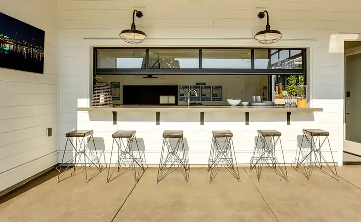 Black-framed aluminum and glass overhead door raised open over an outdoor bar counter, with six wire stools and white shiplap siding.